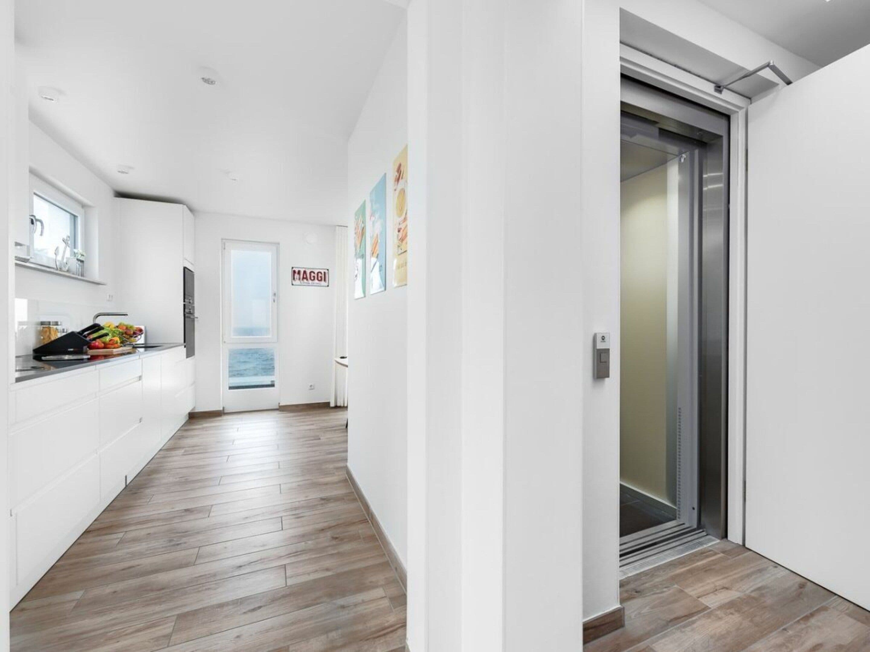 A modern kitchen with white cabinets, a wooden floor, and a view through a door.