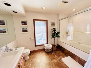A bathroom with a bathtub, a sink, a toilet, and a window with blinds. - Boots & Class, where peaceful Iowa country living meets classy accommodations. (Harlan)