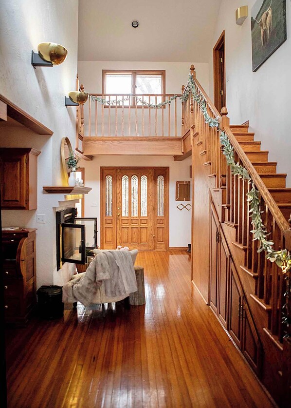 A spacious hallway with a wooden staircase, a dining area with a table and chairs, and a large wooden door with stained glass windows. - Boots & Class, where peaceful Iowa country living meets classy accommodations. (Harlan)