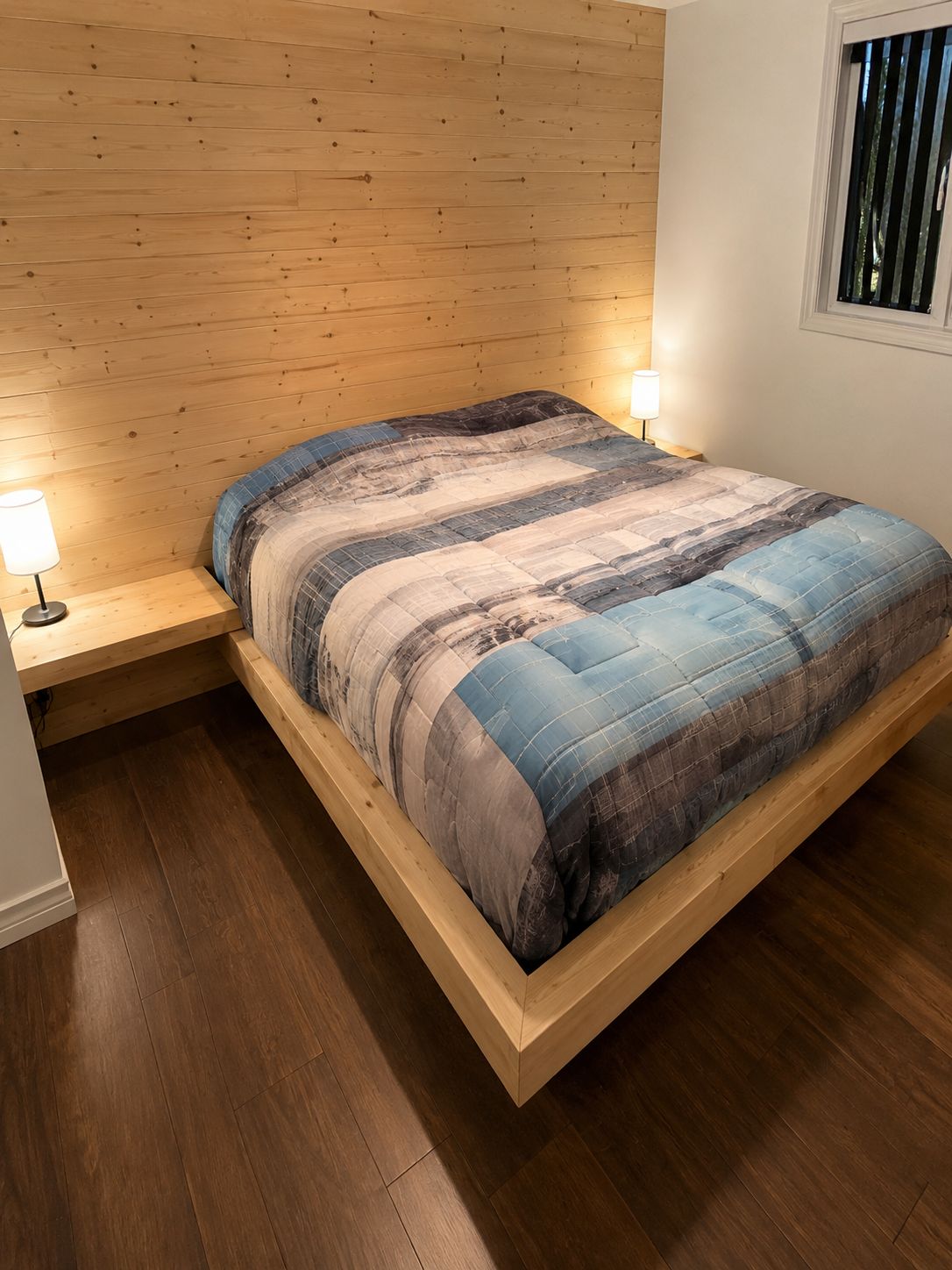 Un lit avec un cadre en bois, une couette bleue et grise, et deux lampes de chevet.