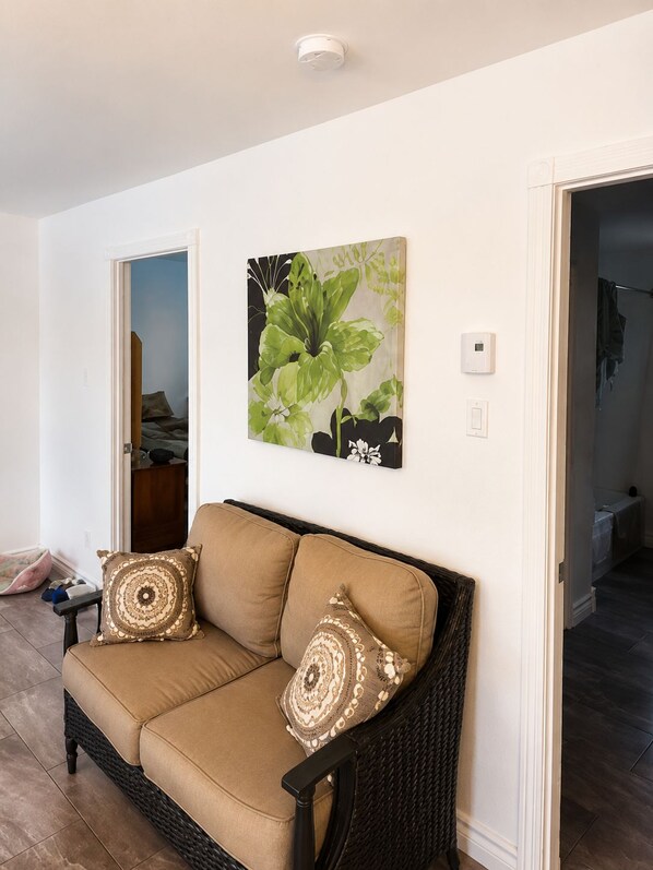 A living room with a beige sofa, two patterned cushions, a wall-mounted artwork of green foliage, and a visible door leading to another room.