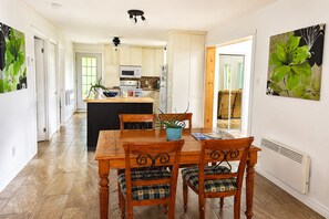 A dining area with a wooden table and chairs, a kitchen in the background, and a large window.