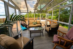 A covered outdoor seating area with wicker furniture, a glass table, and a view of a lake.