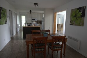 A dining area with a wooden table and chairs, a kitchen in the background, and a view of the outdoors.