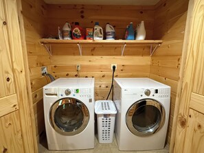 A laundry room with two white front-loading washing machines and a shelf above containing various cleaning products.