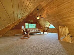 An attic room with a sloped ceiling, a ceiling fan, a sofa, two rocking chairs, and a window with a view of greenery. - Ski-in/Ski-out 9 bed log cabin on Pleasant Mountain with Moose Pond lake access (Bridgton)