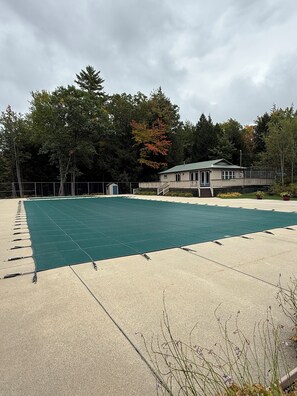 A covered outdoor area with a pool, a small building, and a fenced area with trees in the background.