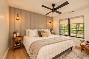 A bedroom with a bed, a ceiling fan, bedside lamps, a window with a view, and a nightstand with decorative items. - 
Walk to Cashiers - 2 king bed rooms + bunk bed with queen, firepit & secluded (Cashiers)