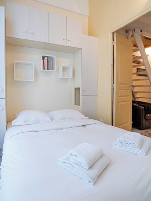 A neatly made bed with white linens and towels, a white wardrobe with open doors, and a staircase leading to an upper level. - Warm Seaside Apartment - Trouville (Trouville-sur-Mer)