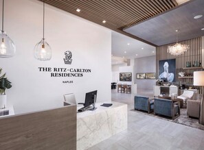 A reception area with a marble counter, a computer, and a desk lamp. There is a reception desk with a logo above it. The area has a modern design with a wooden ceiling and pendant lights. - Stunning Ocean Views 4100 sq ft condo in The Ritz-Carlton Residence (Naples)