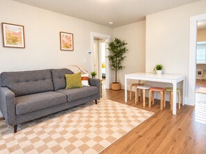 Ein Wohnzimmer mit einer Couch, einem Esstisch mit Hockern, einem gemusterten Teppich und einer Topfpflanze.