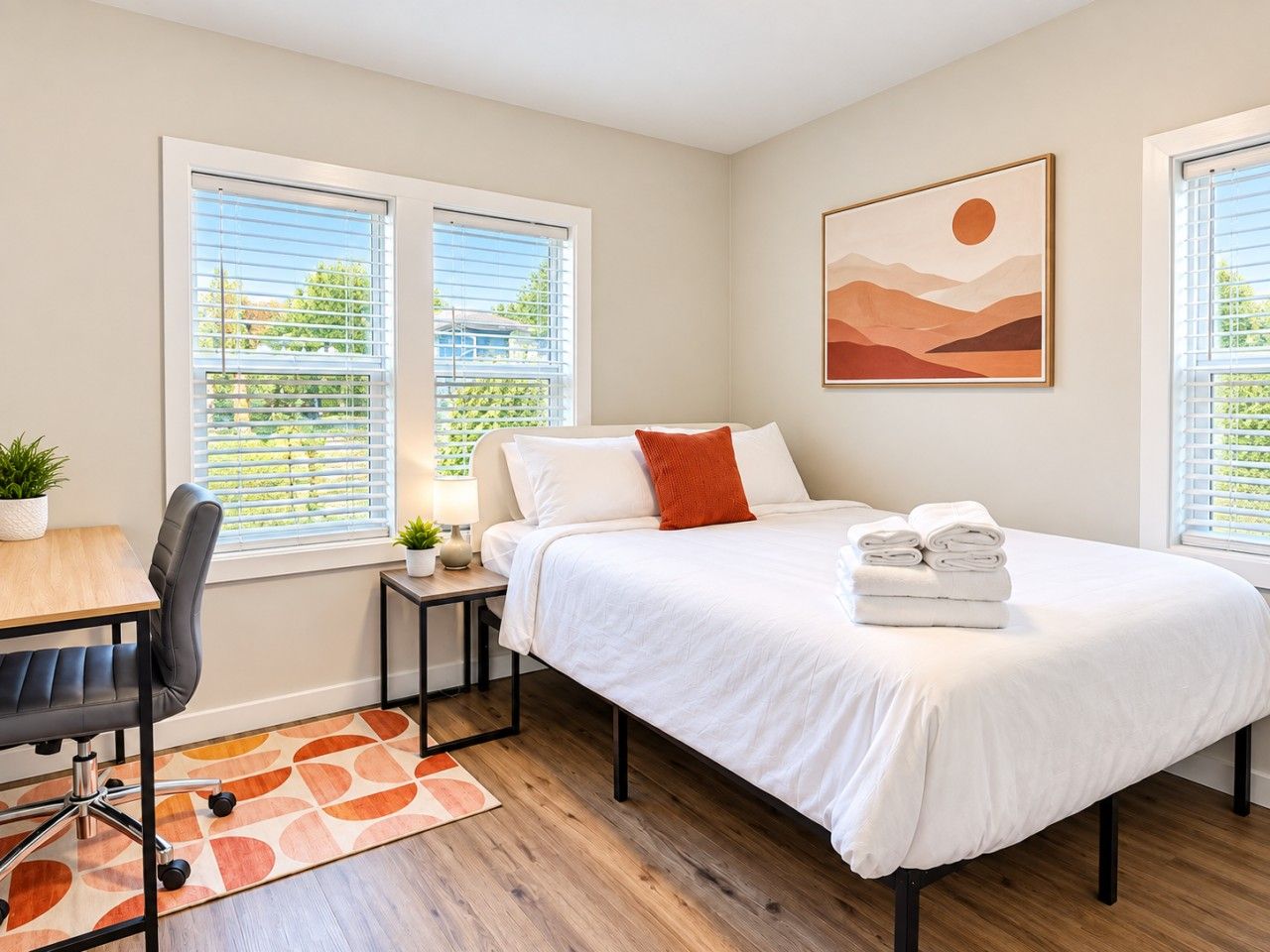 A bedroom with a bed, a desk, and a chair. There are two windows with blinds, a painting on the wall, and a small table with a plant.