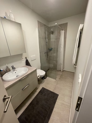 A modern bathroom with a sink, mirror, toilet, shower, and a glass-enclosed shower stall. - Cozy Apartment Near Reykjavík (Hafnarfjordur)