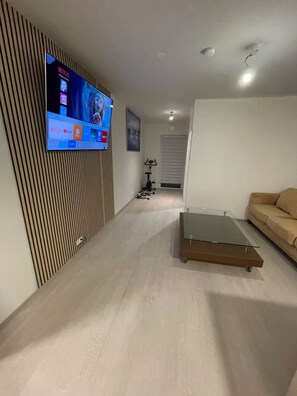 A modern living room with a flat-screen TV mounted on the wall, a glass coffee table, a beige sofa, and a ceiling fan. - Cozy Apartment Near Reykjavík (Hafnarfjordur)