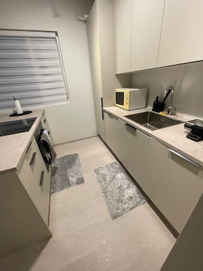 A compact kitchen with a microwave, sink, and a window with blinds. - Cozy Apartment Near Reykjavík (Hafnarfjordur)