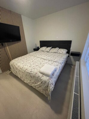 A bedroom with a bed, a flat-screen TV mounted on the wall, a bedside table with a bottle and glasses, and a radiator. - Cozy Apartment Near Reykjavík (Hafnarfjordur)
