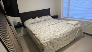 A bedroom with a bed, a nightstand, a window with blinds, and a wall-mounted heater. - Cozy Apartment Near Reykjavík (Hafnarfjordur)