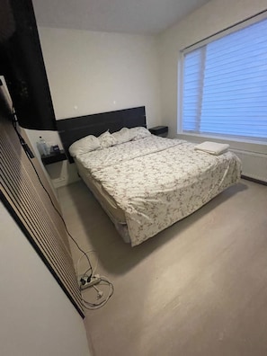 A bedroom with a bed, a nightstand, a window with blinds, and a wall-mounted heater. - Cozy Apartment Near Reykjavík (Hafnarfjordur)