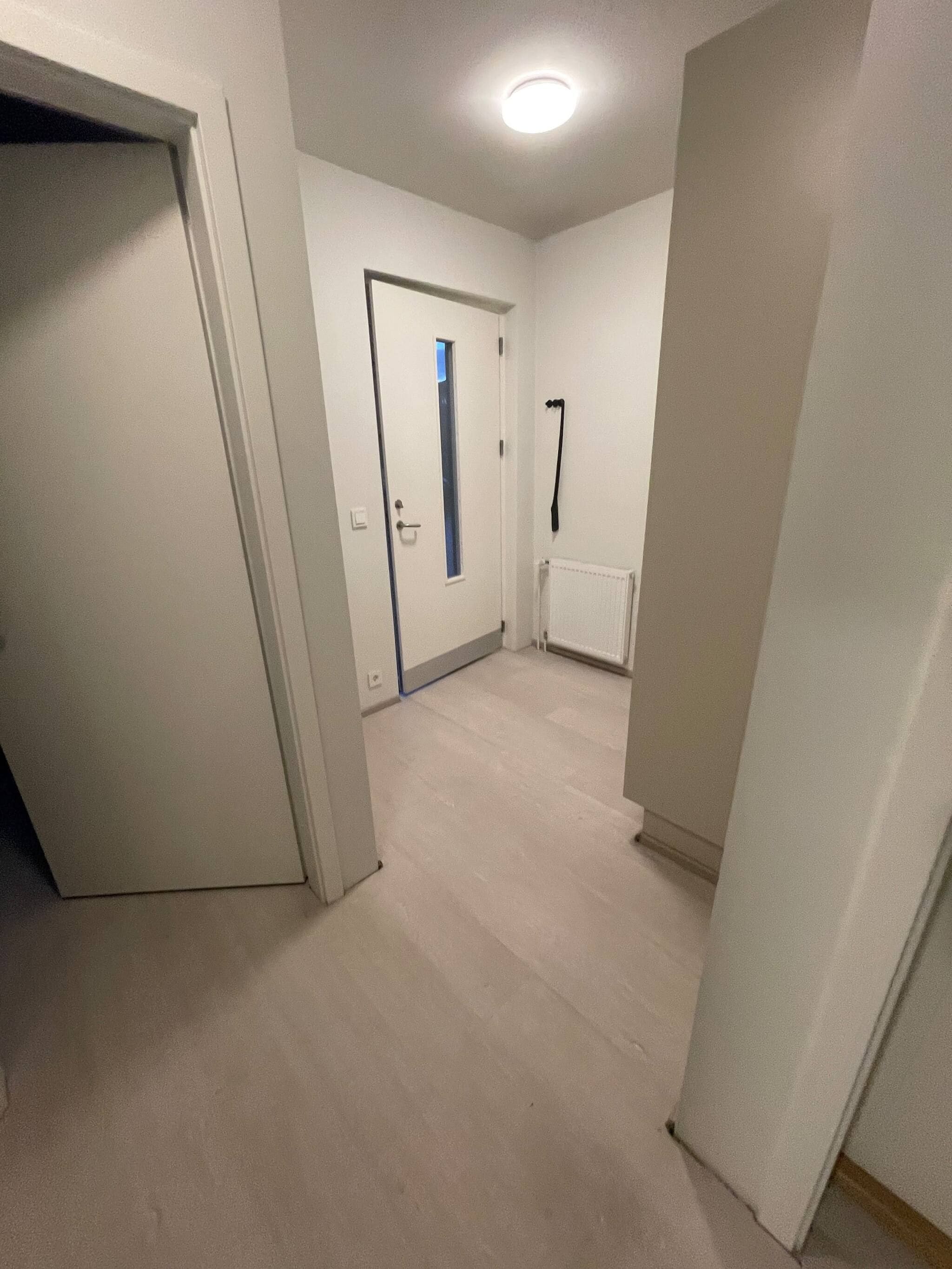 Un pasillo con una puerta que da a una habitación, un radiador y una ventana.