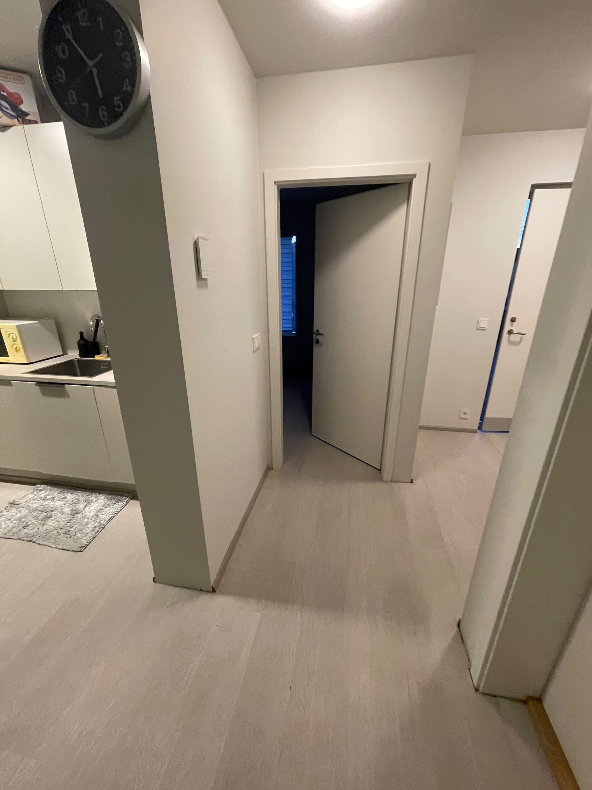 Un pasillo estrecho con una puerta que da a una habitación oscura, un reloj en la pared y una pequeña cocina con microondas y fregadero.