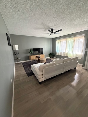 Sala de estar com sofá, televisão, ventilador de teto e janela com cortinas.