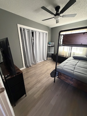 Quarto com beliche, ventilador de teto, televisão sobre suporte e janela com cortinas.