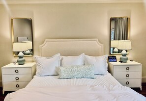 A hotel room with a large bed, two bedside tables with lamps, a mirror, and a small table with a vase and a heart-shaped decoration. -  Lovely house within walking distance to TCU (Fort Worth)