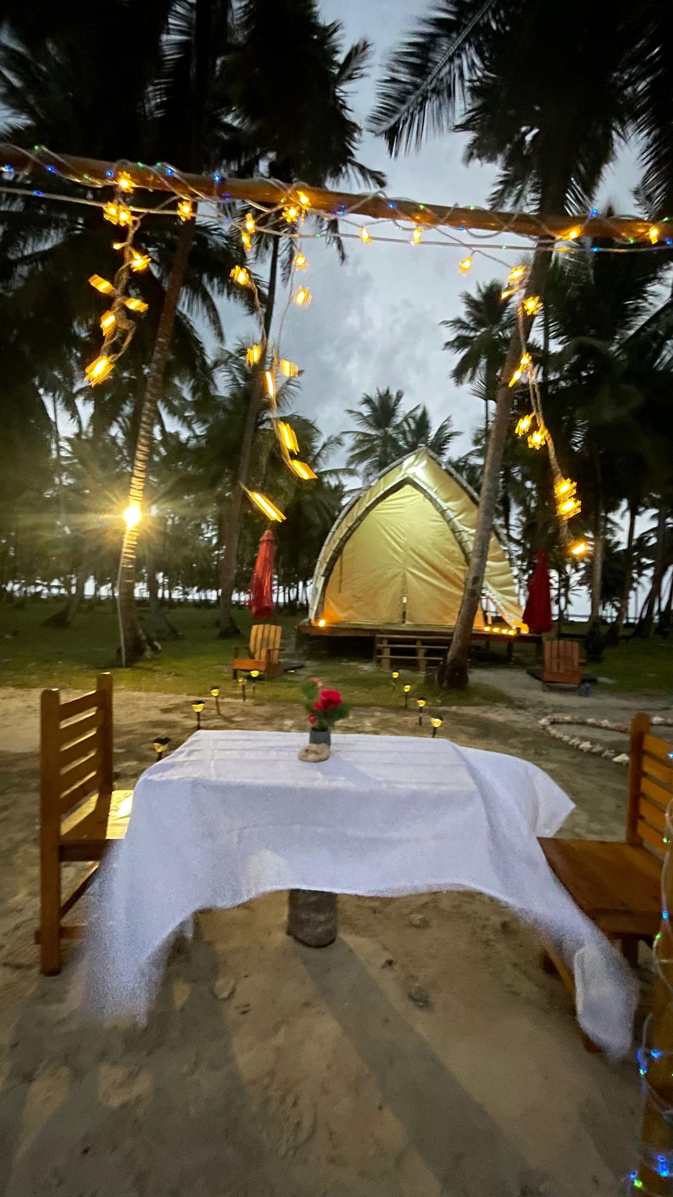 A tent with a table and chairs set up on a sandy area, surrounded by palm trees and string lights.