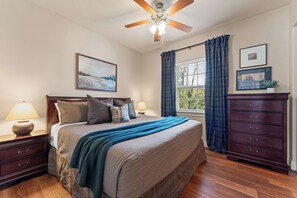 Ein Schlafzimmer mit Bett, Nachttischen, Kommode, Deckenventilator, Fenster mit Vorhängen und Bildern an den Wänden.