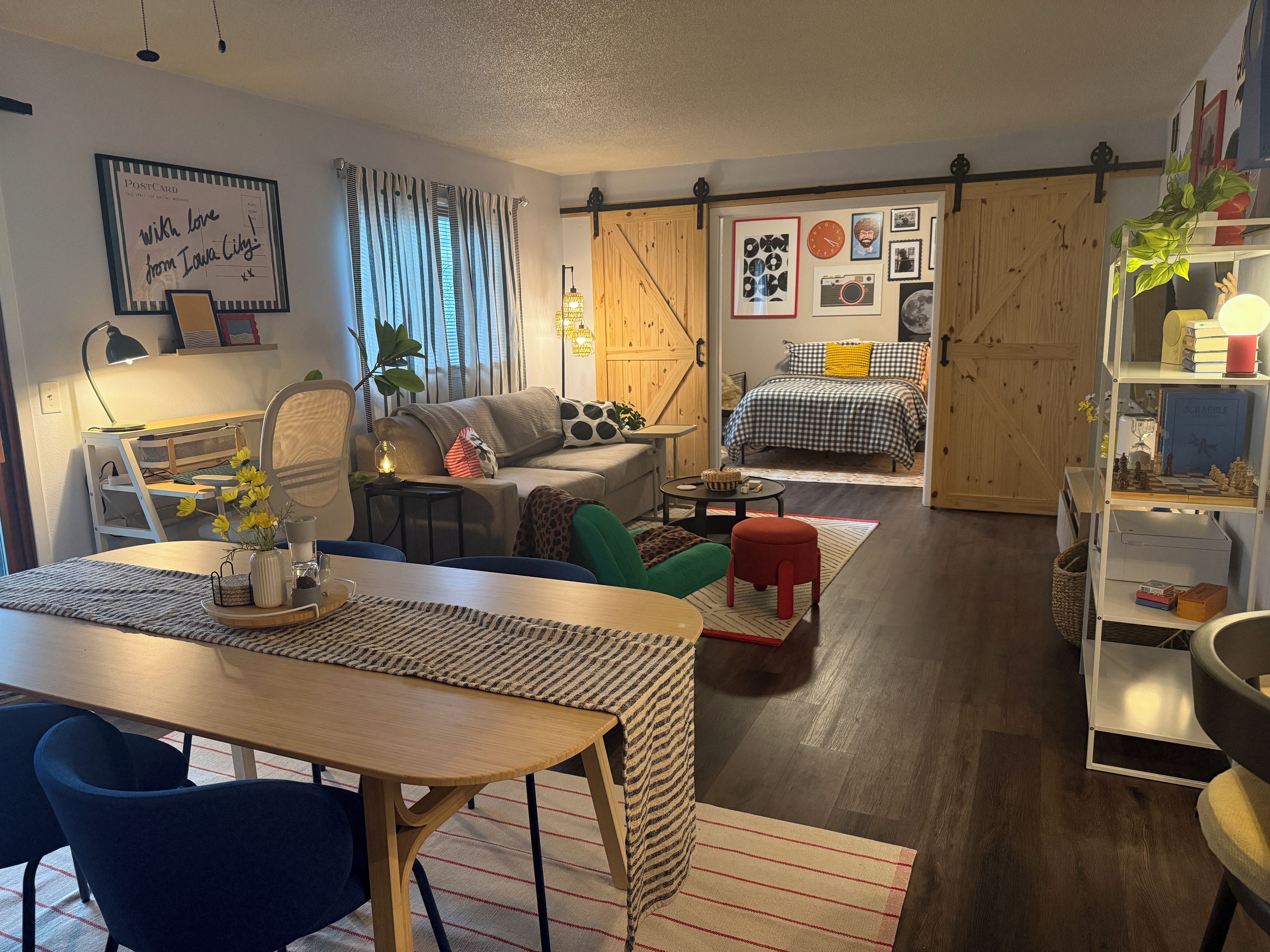 A cozy living room with a dining area, a bed with checkered bedding, and a bookshelf with various items.