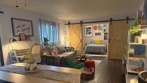 A cozy living room with a dining area, a bed with checkered bedding, and a bookshelf with various items.