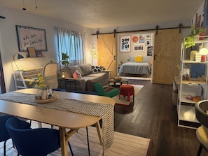 Un salon chaleureux avec un coin repas, un lit avec une couette à carreaux et une bibliothèque remplie de divers objets.