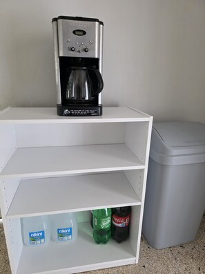 Ein weißes Regal mit einer Cuisinart Kaffeemaschine obenauf, zwei leeren Regalböden darunter und einem Mülleimer zur Rechten.