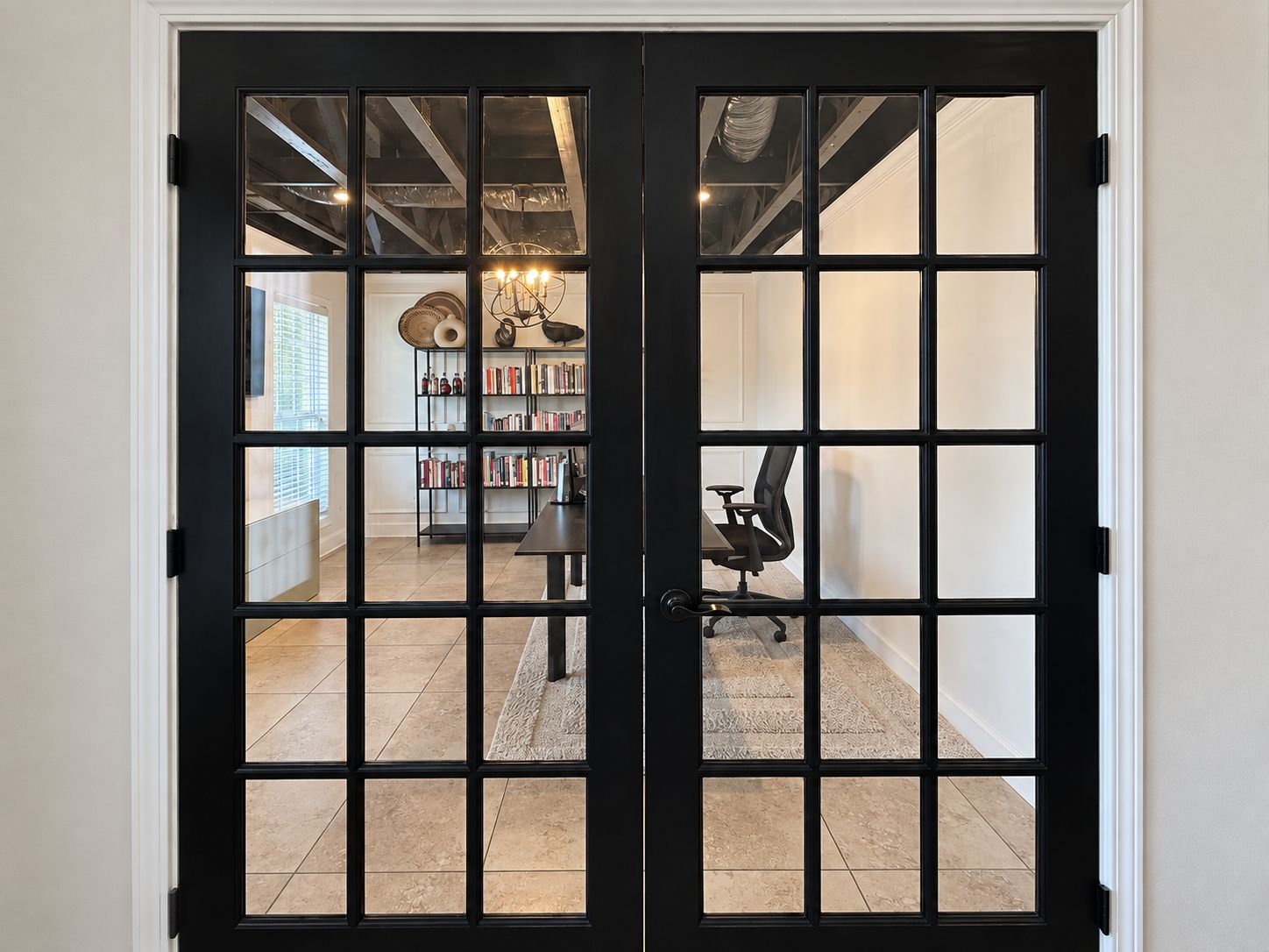 Una puerta con marco negro y diseño de cuadrícula, que da paso a una habitación con una estantería, un escritorio y una silla.