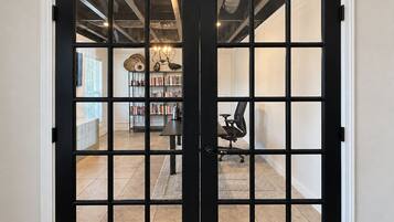 Una puerta con marco negro y diseño de cuadrícula, que da paso a una habitación con una estantería, un escritorio y una silla.