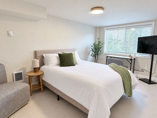 A modern hotel room with a bed, a side table, a lamp, a television, and a window with blinds.