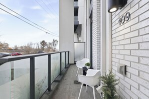 Un balcon doté d’une balustrade en verre, deux chaises blanches, une petite plante en pot et un immeuble à la façade en briques blanches, portant le numéro 109.