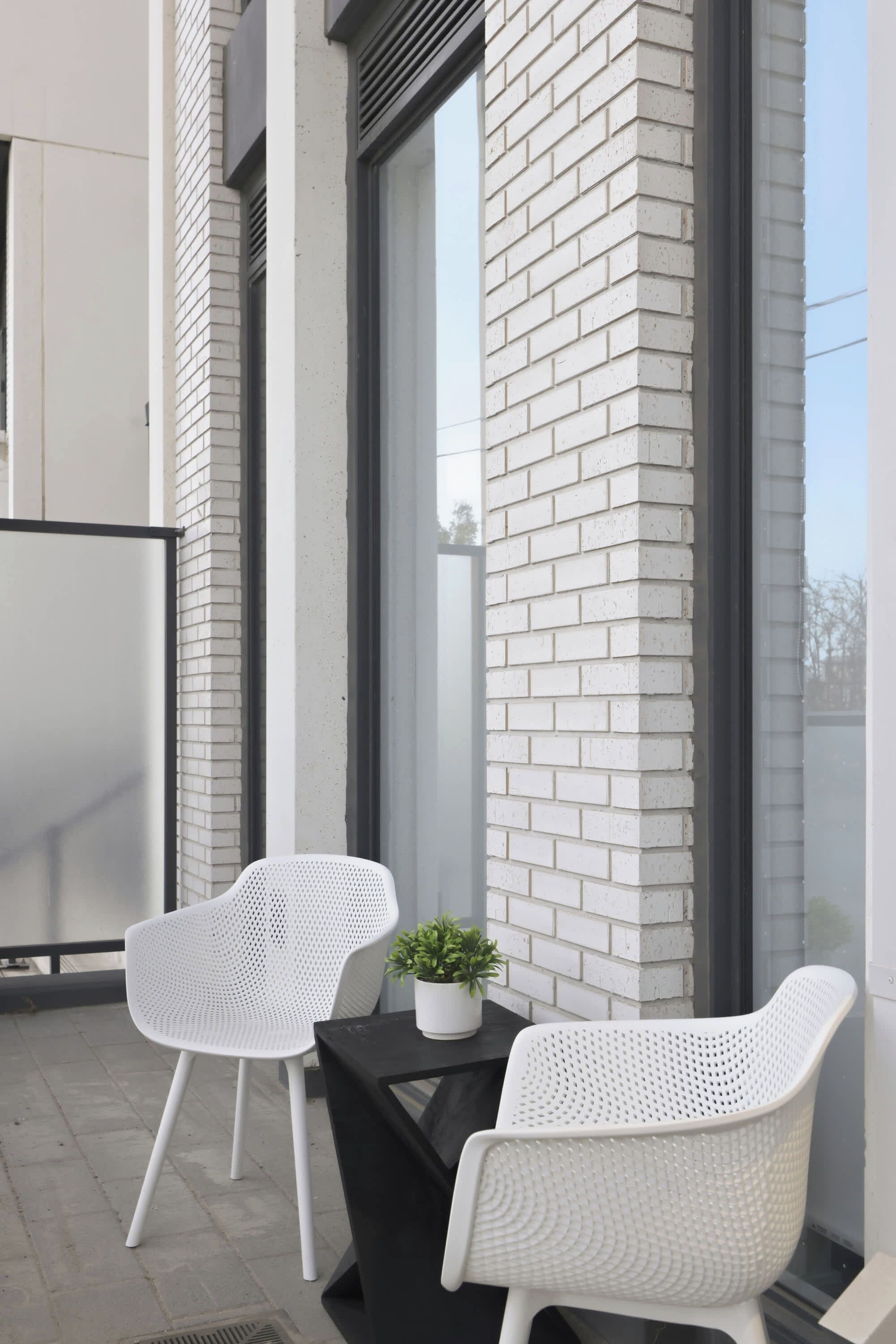 A balcony with two white wicker chairs, a small table with a potted plant, and a large window.