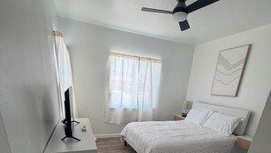 Un dormitorio con cama, escritorio, ventilador de techo y ventana con cortinas.