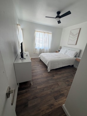 Ein Schlafzimmer mit einem Bett, einem Schreibtisch, einem Deckenventilator und einem Fenster mit Vorhängen.