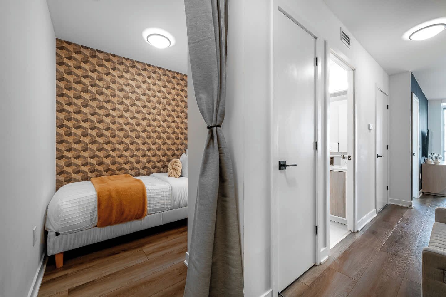 A modern bedroom with a geometric-patterned headboard, a bed with a white comforter and an orange throw blanket, and a wooden floor.