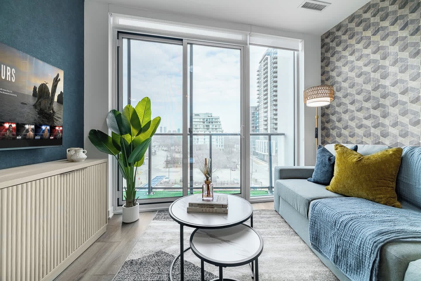 A modern living room with a sofa, a coffee table, a plant, and a large window with a city view.