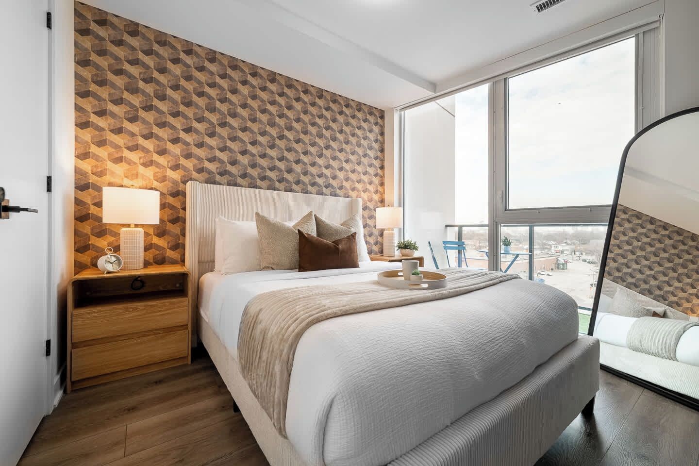 A modern hotel room with a large bed, a nightstand with a lamp, a bedside table with a clock, and a view of the city through large windows.