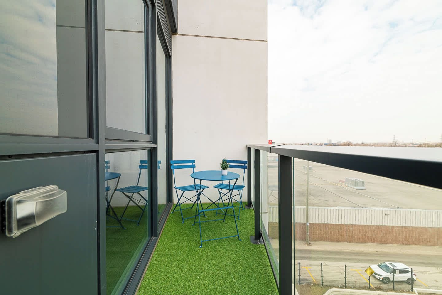 A balcony with a glass door, a small table with blue chairs, and a view of an airport.