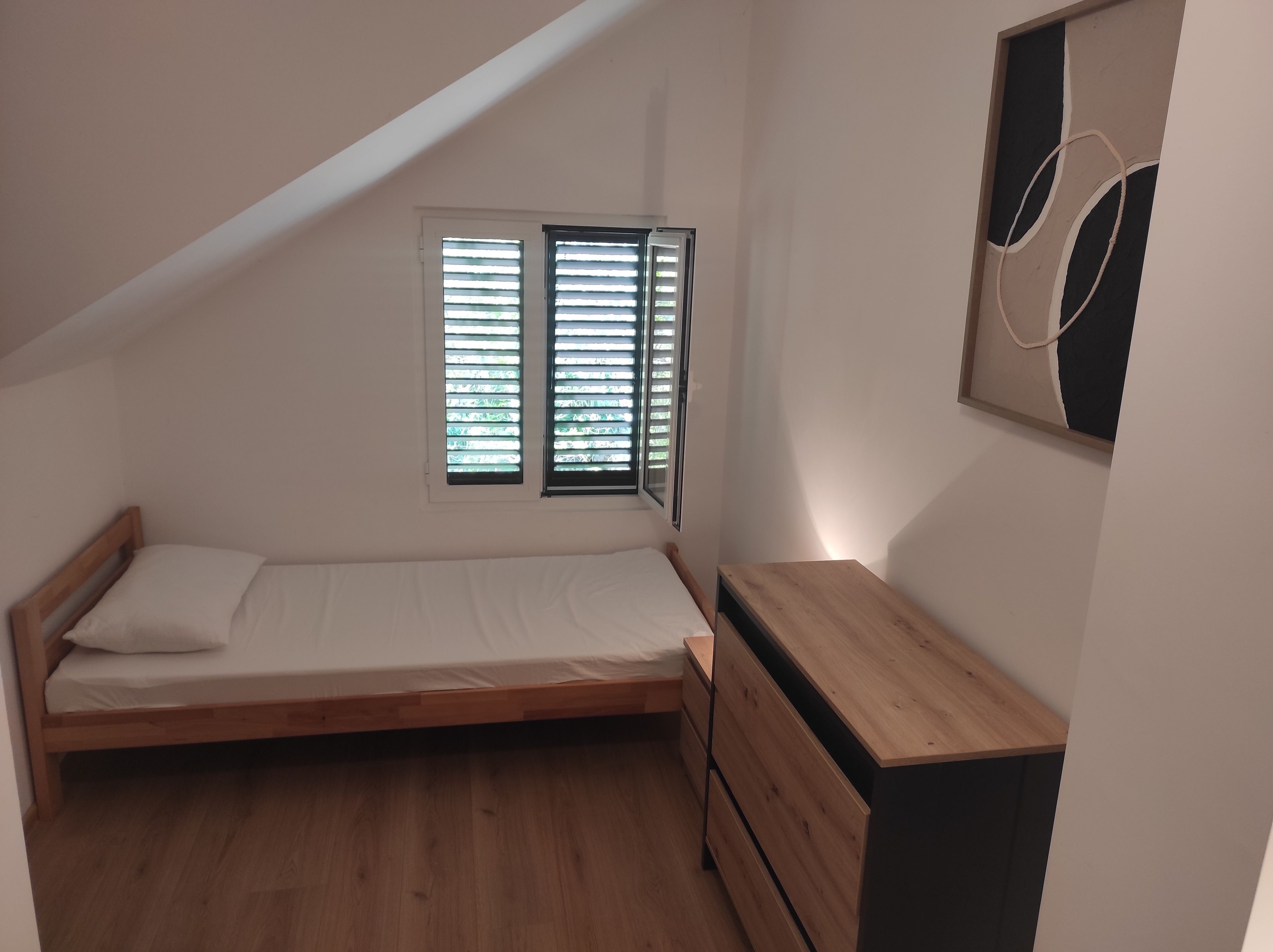Une petite chambre avec un lit simple, un plafond en pente, une commode en bois et une fenêtre avec des stores.