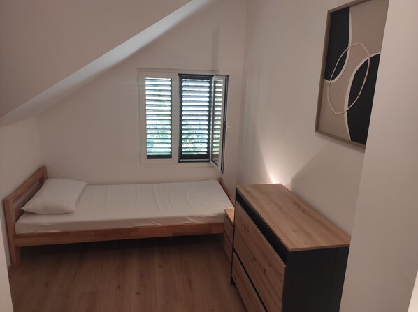 A small, single-bed room with a sloped ceiling, a wooden dresser, and a window with blinds.