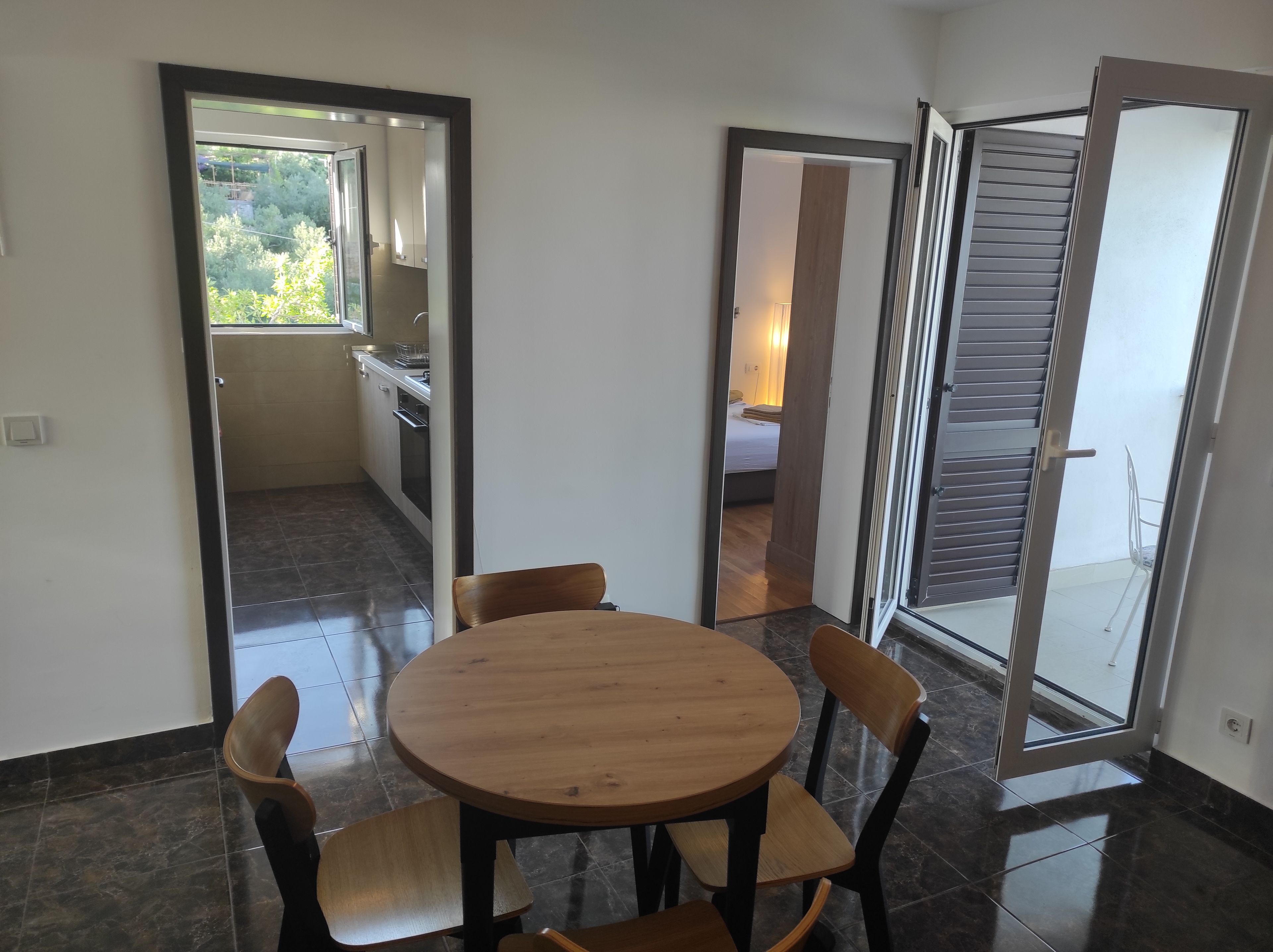 Une pièce avec une table ronde en bois et quatre chaises, une porte-fenêtre coulissante donnant sur un balcon, et une vue sur la cuisine et la chambre à coucher à travers une porte ouverte.