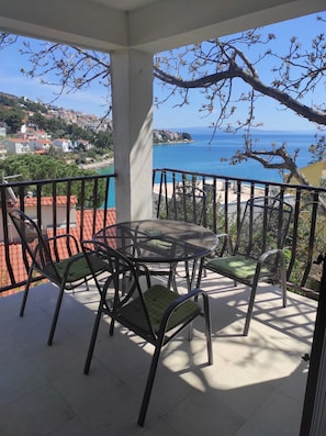 Un balcón con mesa y sillas, con vista al mar y edificios bajo un cielo despejado.
