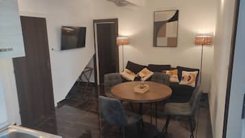 A modern dining area with a round table, chairs, and a sofa with cushions. There is a TV mounted on the wall and a floor lamp.