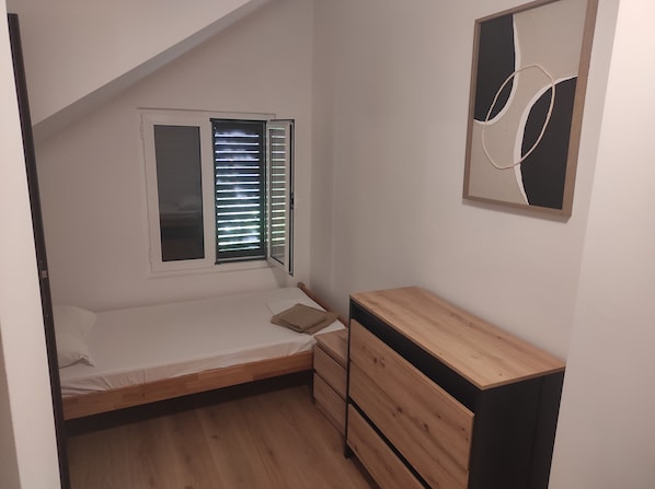 A small, single-bed room with a wooden headboard, a bedside table, and a dresser. There is a window with blinds, and a framed abstract artwork on the wall.